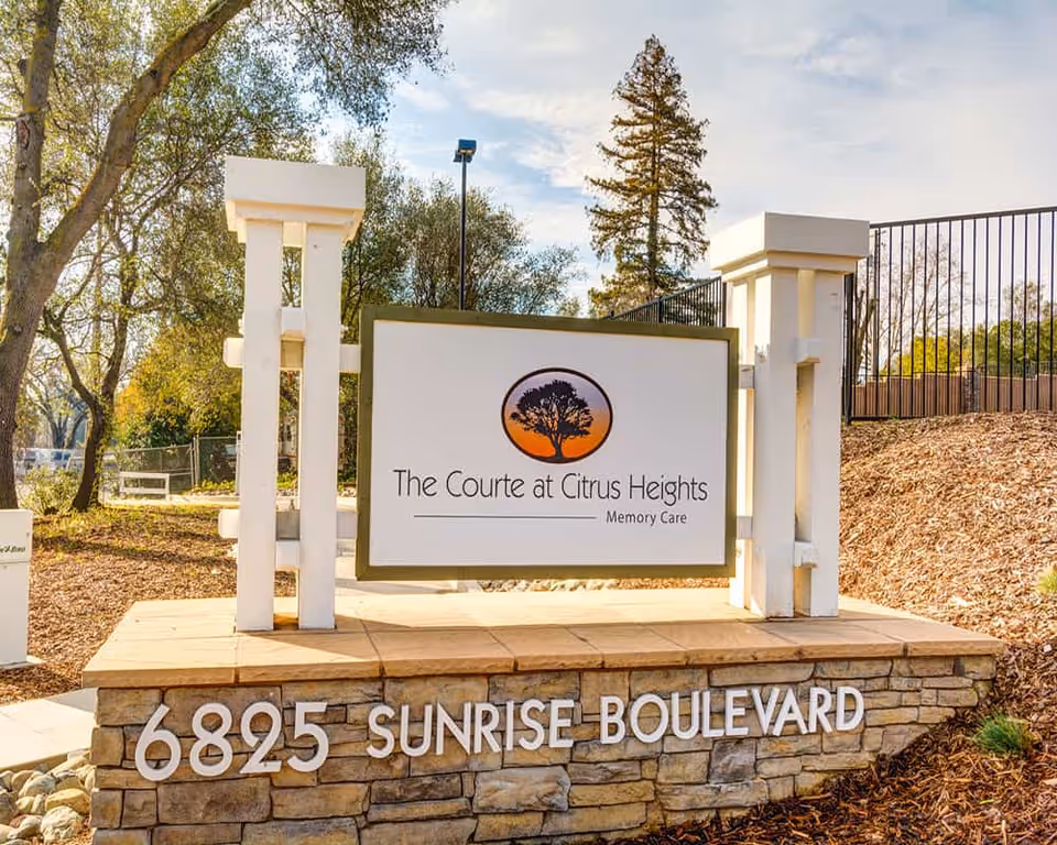 Outdoor sign for The Courte at Citrus Heights Memory Care facility, mounted on a stone base with the address 6825 Sunrise Boulevard, surrounded by trees and landscaping.