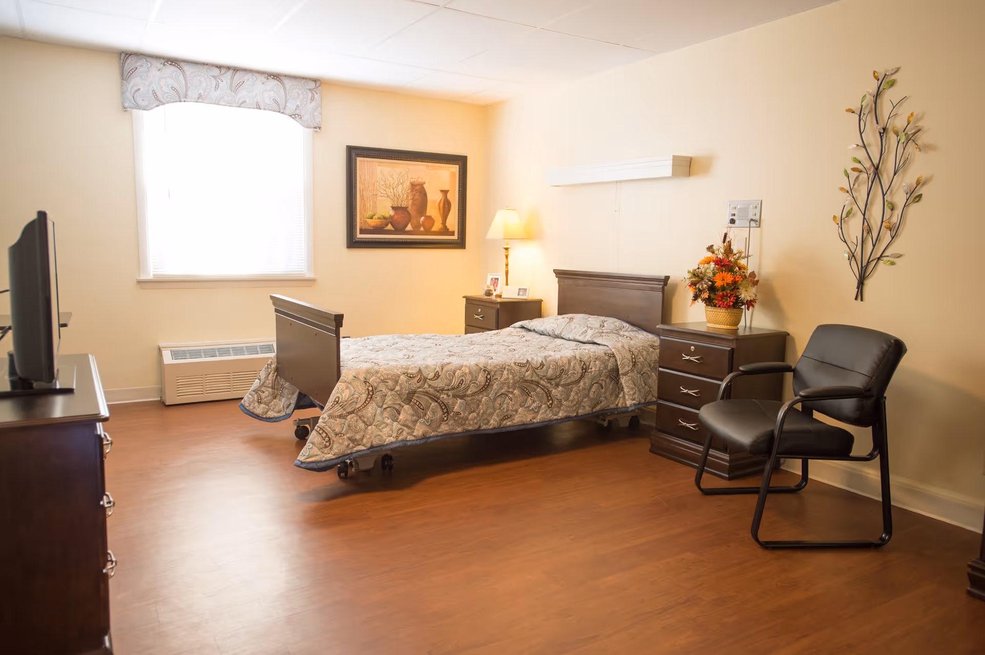 A furnished nursing facility bedroom with a single bed, nightstand, armchair, and a television.