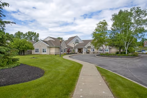 Exterior view of a single-story senior living facility building with beige siding and stone accents, surrounded by green lawns, trees, and a curved driveway under a partly cloudy sky.