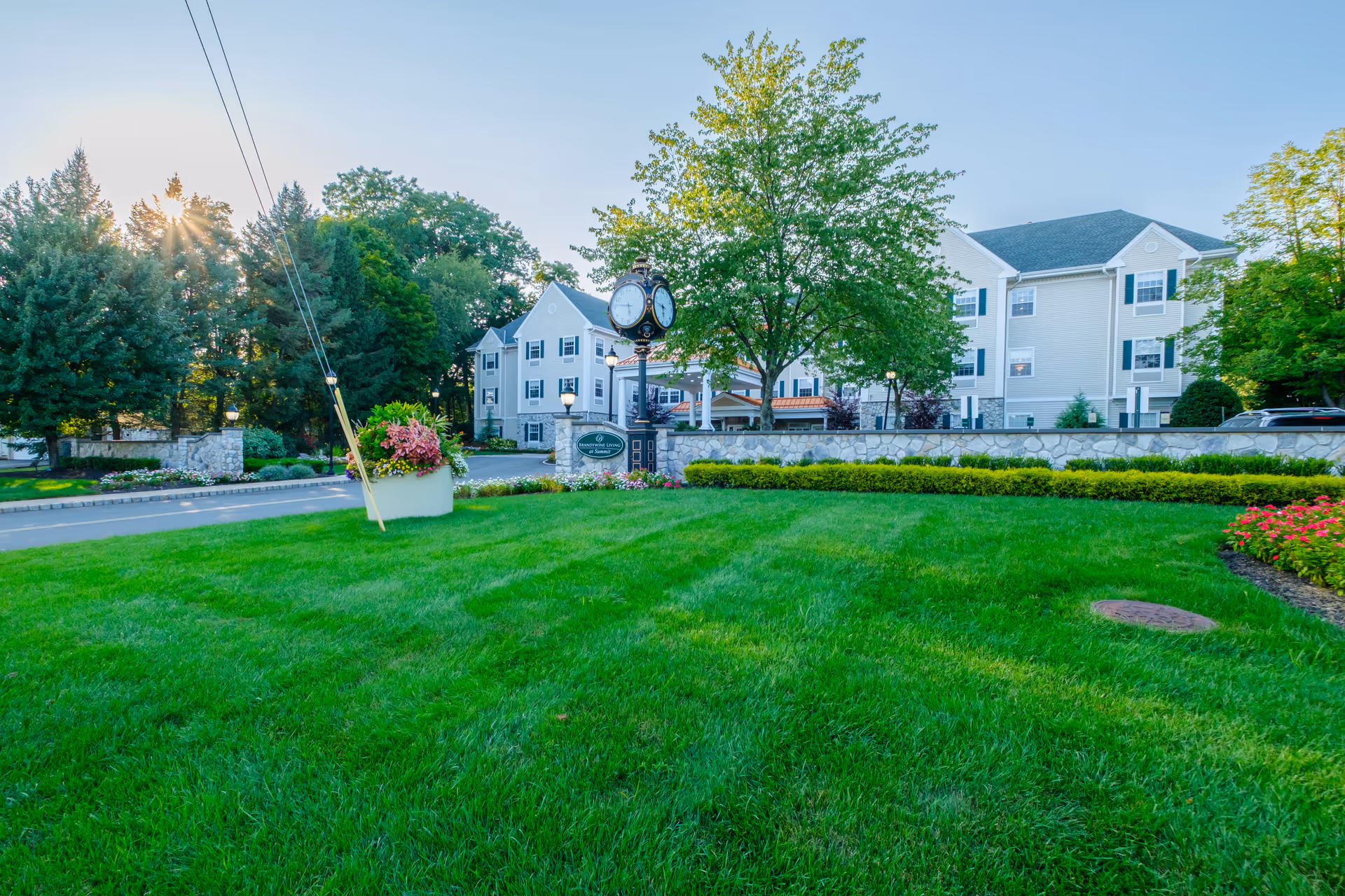 Exterior view of Brandywine Summit by Monarch facility with a large green lawn in the foreground, a decorative clock on a post, trees, and a multi-story building with white siding and dark shutters in the background under a clear sky.