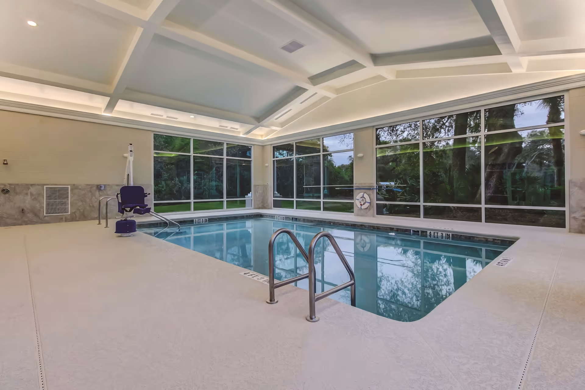 Indoor swimming pool area with large windows showing greenery outside. The pool has metal handrails and a pool lift chair for accessibility. The ceiling features recessed lighting and a coffered design.
