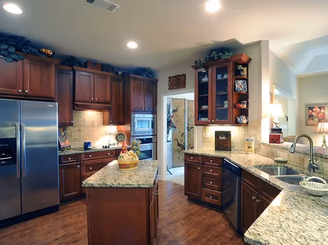 A spacious kitchen with wooden cabinets, granite countertops, and stainless steel appliances including a refrigerator and built-in oven. There is a kitchen island in the center with a decorative ceramic container on top. The kitchen has recessed lighting and a double sink with a modern faucet. The floor is wooden, and there are decorative items on top of the cabinets and shelves.