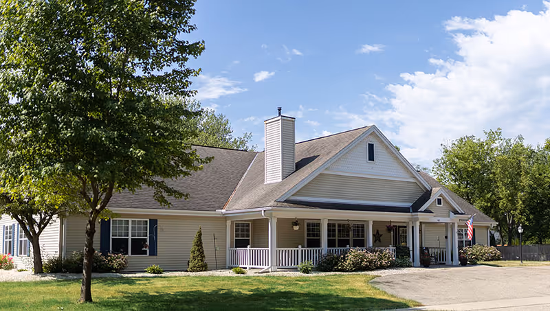 Single-story beige senior living building with a front porch, landscaped shrubs, trees, and an American flag under a blue sky.