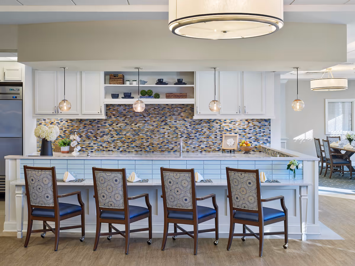 Bright communal kitchen and breakfast bar with four upholstered chairs, mosaic tile backsplash, pendant lights, and a dining area in the background.