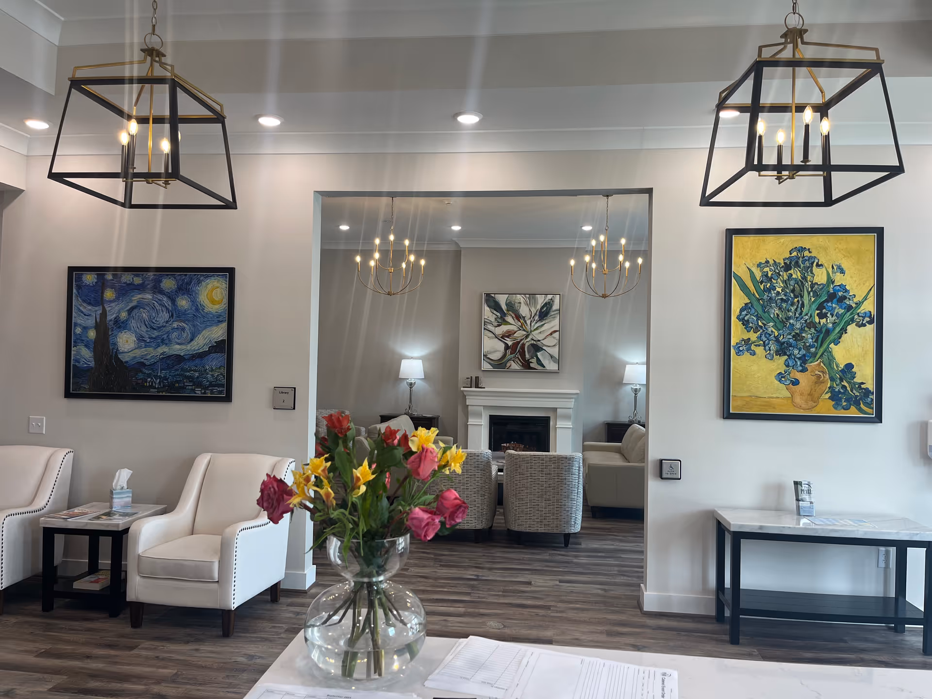 A bright and modern living room area with white walls and wooden flooring. The foreground features a glass vase with colorful flowers on a white table with papers. Two white armchairs are positioned near a small black side table with tissues and magazines. The background shows a cozy seating area with a fireplace, two patterned armchairs, a beige sofa, and two table lamps. Two large framed paintings hang on the walls, one depicting Van Gogh's Starry Night and the other a vase with blue flowers. Two geometric pendant lights hang from the ceiling.