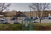 Two-story brick senior living building with a parking lot and a green bus stop shelter.