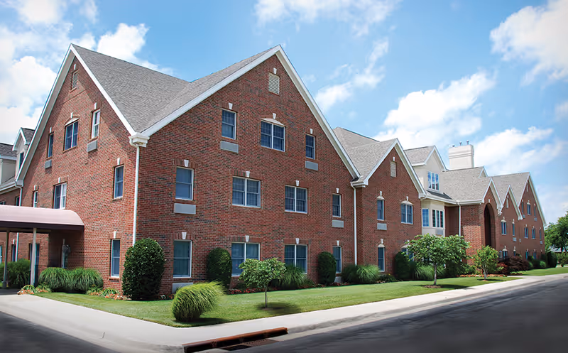 Exterior view of a large brick building with multiple windows and a steep gray roof under a partly cloudy blue sky. The building is surrounded by neatly trimmed bushes, small trees, and green grass along a paved road.