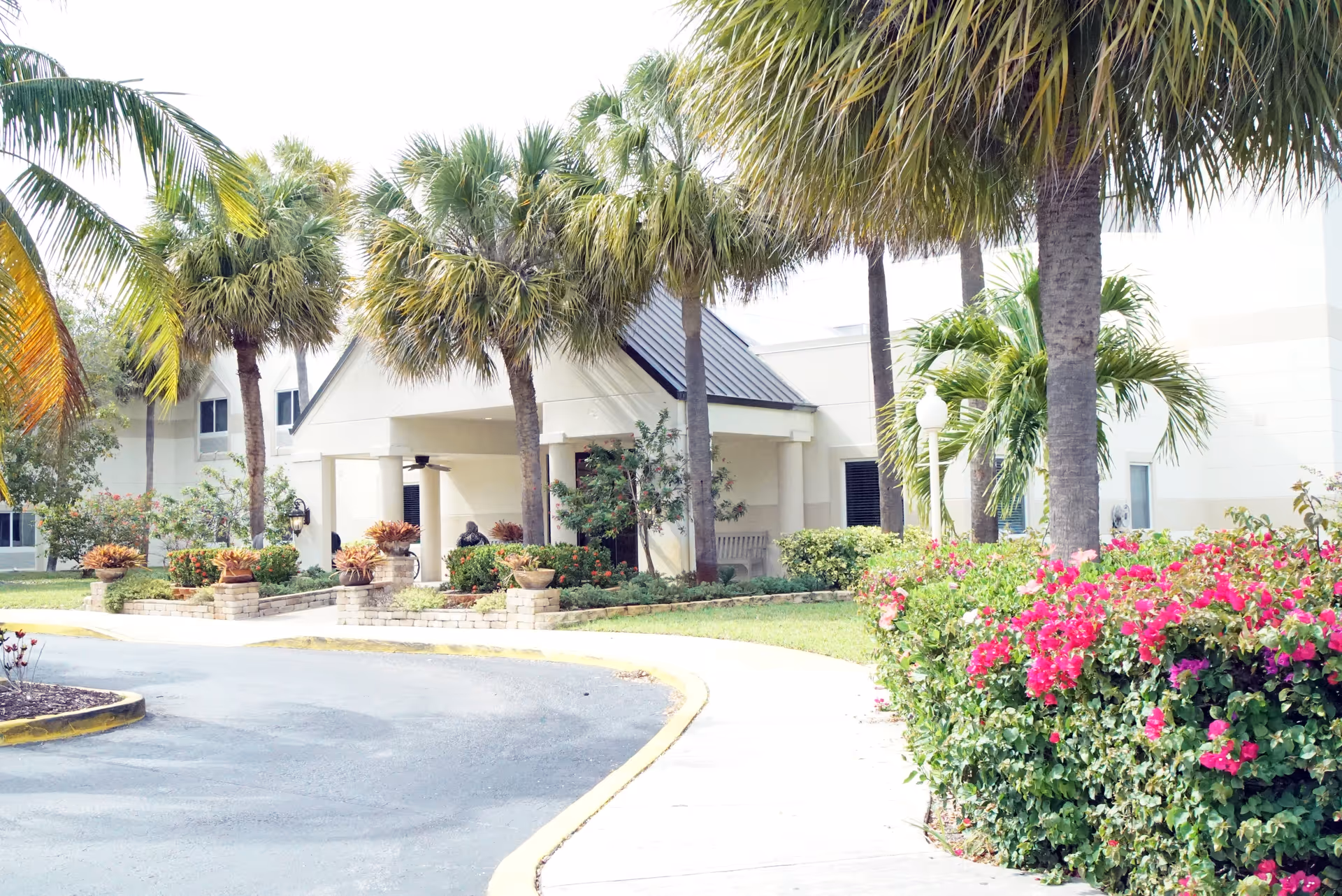 Exterior view of Palm Garden of Aventura facility showing a driveway, landscaped garden with palm trees and flowering bushes, and the entrance to the building with a covered porch.