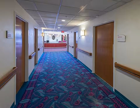 A well-lit hallway with patterned blue and purple carpet, wooden doors on both sides, handrails along the walls, and a reception desk visible at the end of the corridor.