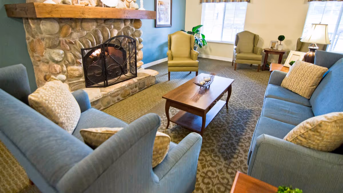 A cozy living room with two blue sofas facing each other, a wooden coffee table in the center, and a stone fireplace with a fire burning. There are two green armchairs near the window, a side table with a lamp, and decorative items on the mantel and tables.