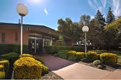 Exterior view of a single-story building with a covered entrance, surrounded by well-maintained bushes, trees, and a paved walkway under a clear blue sky.
