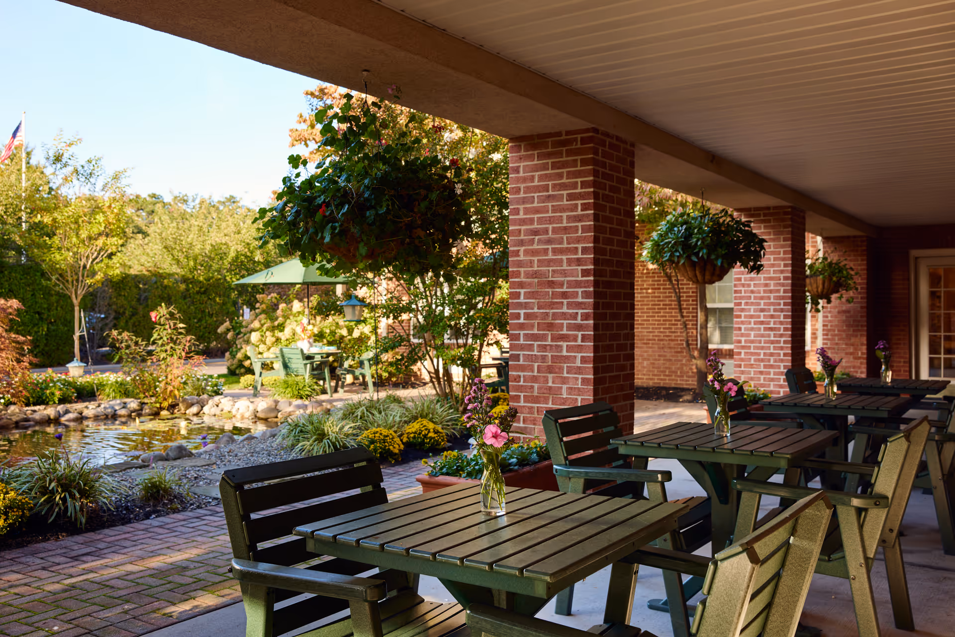 Outdoor covered patio area with green wooden tables and chairs, each table decorated with a small vase of flowers. Hanging flower baskets are suspended from the ceiling. In the background, there is a garden with trees, shrubs, and a small pond surrounded by rocks. A green umbrella shades a seating area further back.