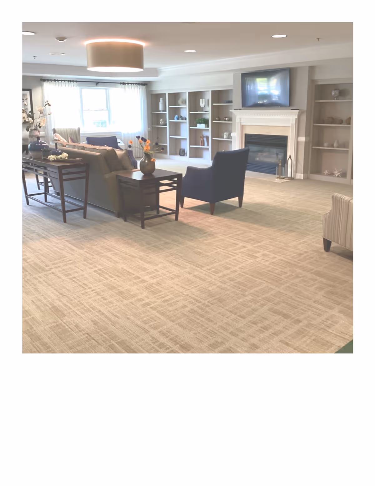 A spacious living room area with beige carpet flooring, a beige sofa, a dark armchair, and a side table with a vase of flowers. The room features built-in shelves with decorative items, a fireplace with a mounted TV above it, and large windows with sheer curtains allowing natural light to fill the space.