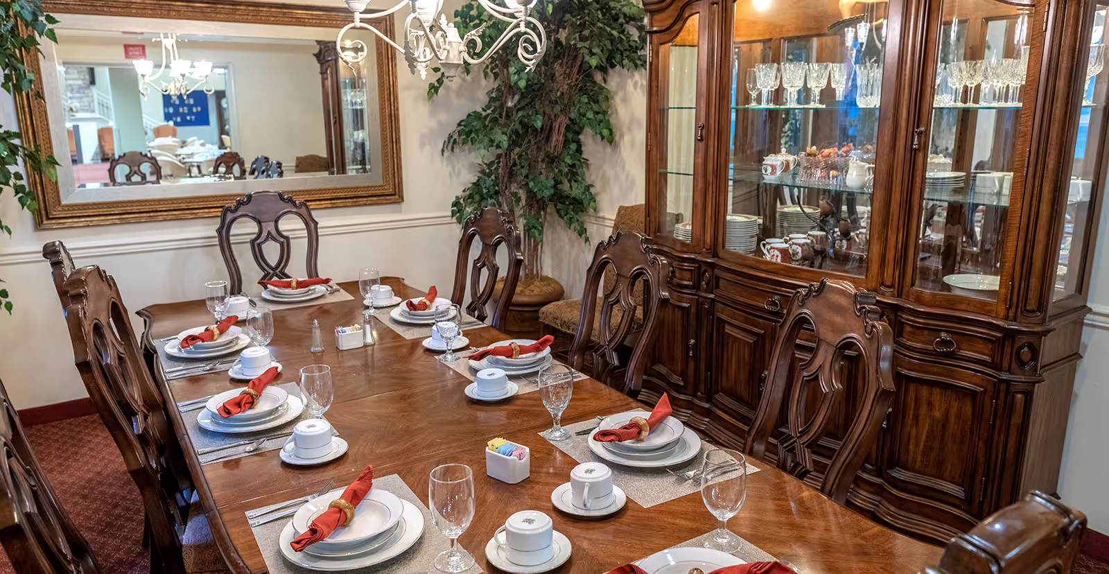 A formal dining room with a long wooden table set for eight people. Each place setting includes a white plate, a cup and saucer, a water glass, silverware, and a red cloth napkin with a napkin ring. Behind the table is a large wooden china cabinet filled with glassware and dishes. A large mirror hangs on the wall reflecting another dining area, and there is a chandelier above the table. A potted plant is in the corner of the room.
