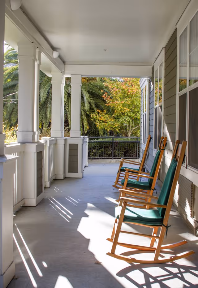 A covered outdoor porch area with three wooden rocking chairs with green cushions lined up along the right side. White pillars and railings border the porch, and lush green trees and palm fronds are visible in the background.
