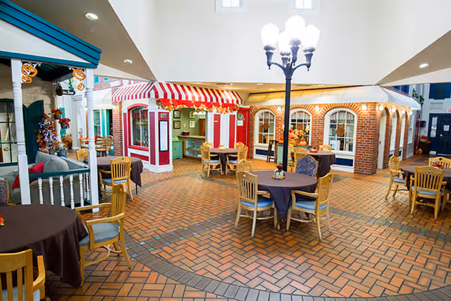 Indoor common area designed like a small village square with brick flooring, round tables covered with dark tablecloths, wooden chairs, a streetlamp-style light fixture, and storefront facades with striped awnings and windows.