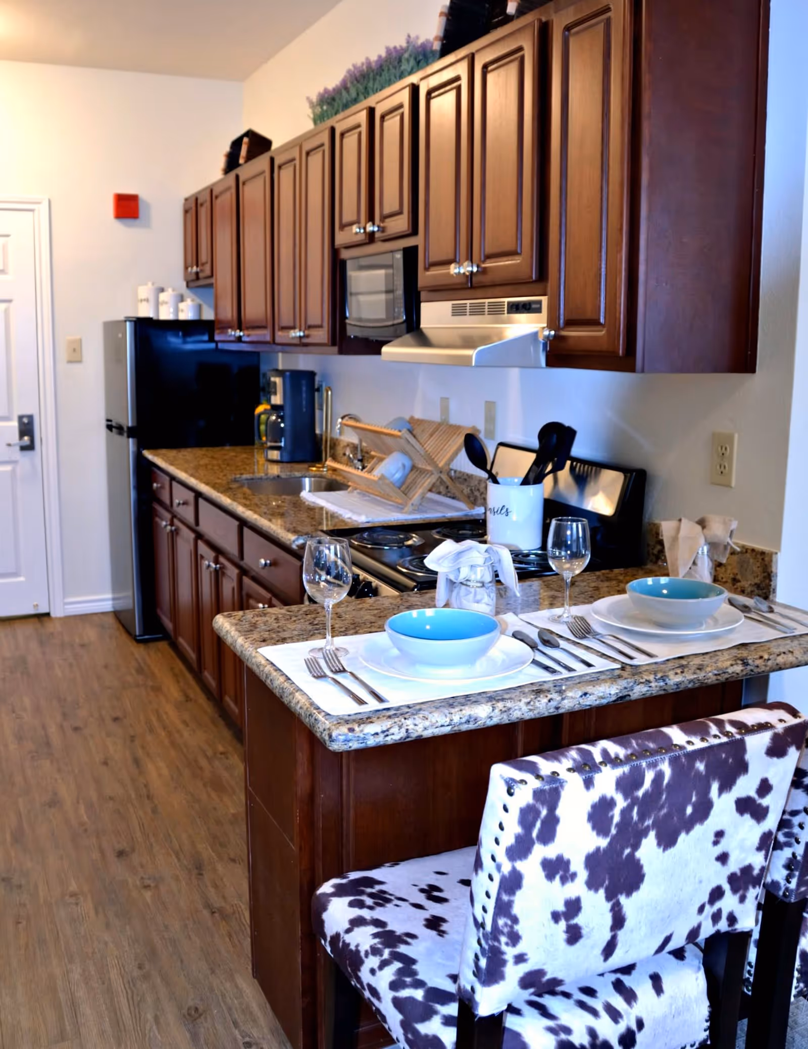 A modern kitchen with dark wooden cabinets, granite countertops, and a small breakfast bar set for two with blue bowls, white plates, silverware, and wine glasses. The kitchen includes a black refrigerator, a microwave, a stove with a range hood, a coffee maker, and a dish drying rack. The floor is wooden, and there are two chairs with a black and white cowhide pattern at the breakfast bar.