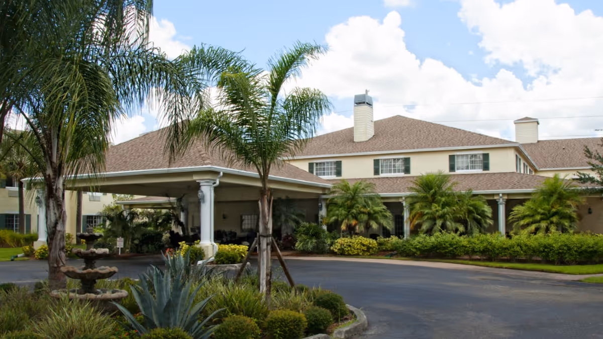 Exterior view of Lexington Manor senior living facility showing a two-story building with a covered entrance, surrounded by palm trees, shrubs, and a circular driveway under a partly cloudy sky.