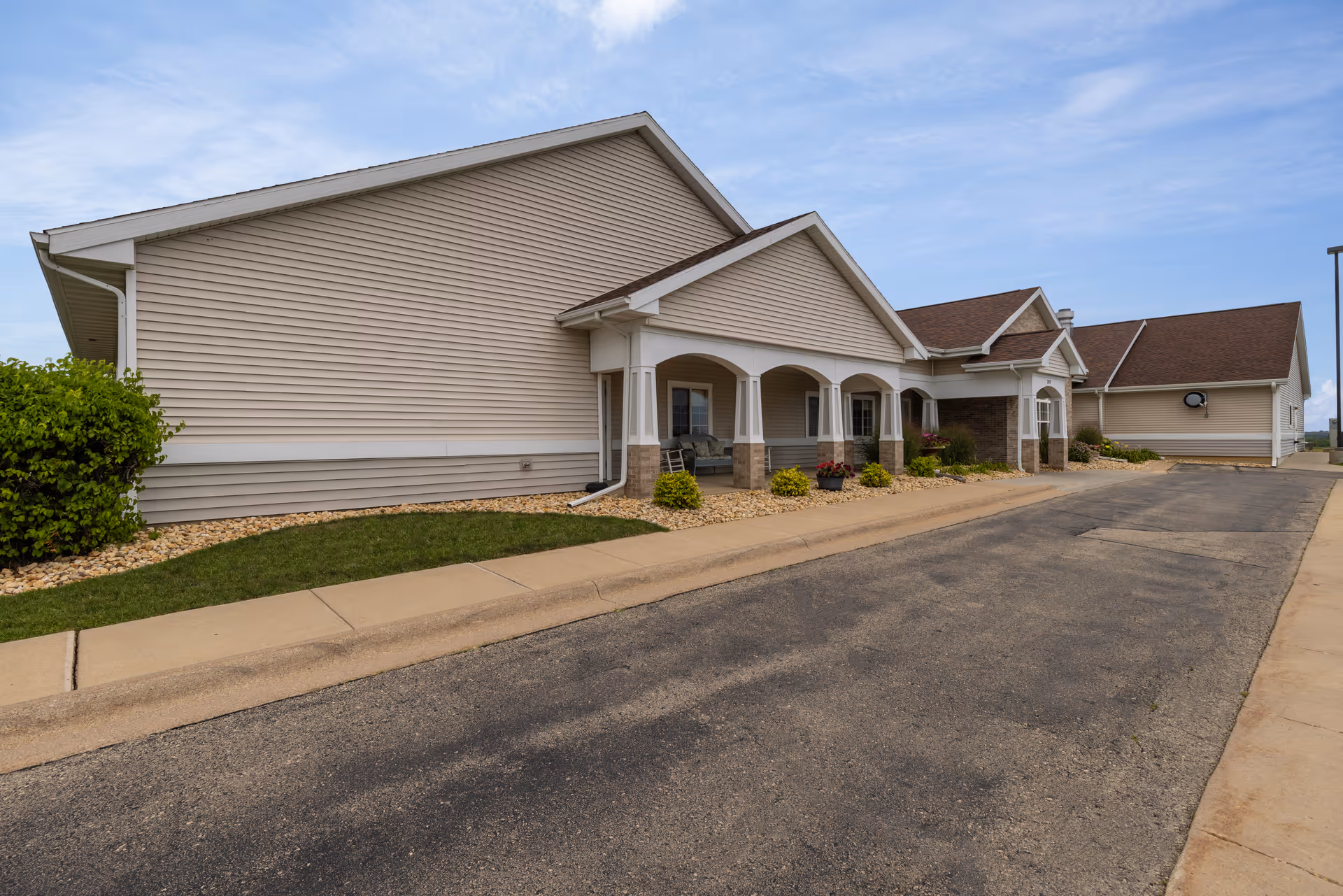 Exterior view of a single-story building with beige siding and a brown roof, featuring a covered porch with white columns and some landscaping with bushes and flowers along the sidewalk.