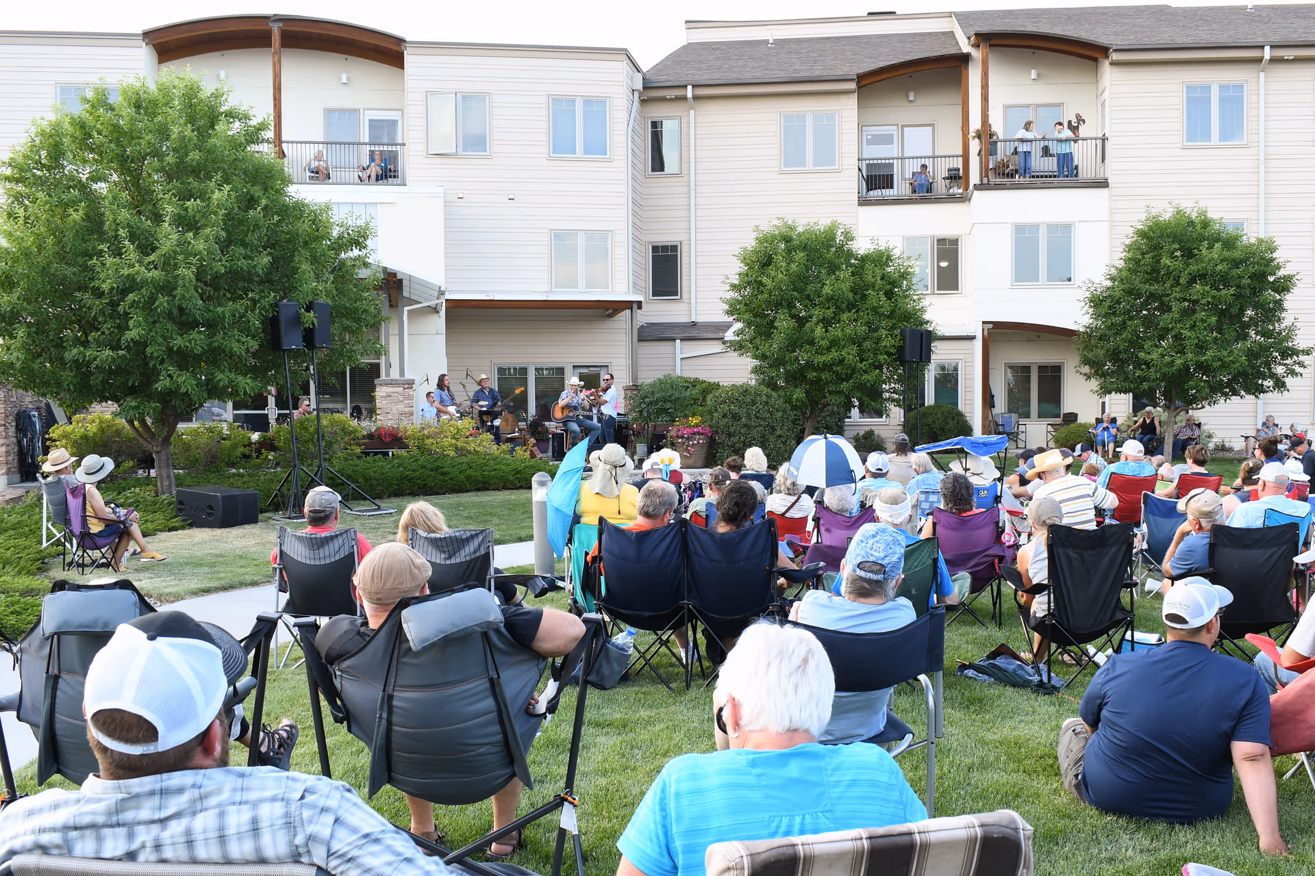 A large group of people seated on lawn chairs and blankets on a grassy area, watching a live band perform outside in front of a multi-story residential building with balconies. The scene is set in a well-maintained garden with trees and shrubs, and some people are standing or sitting on the balconies watching the event.
