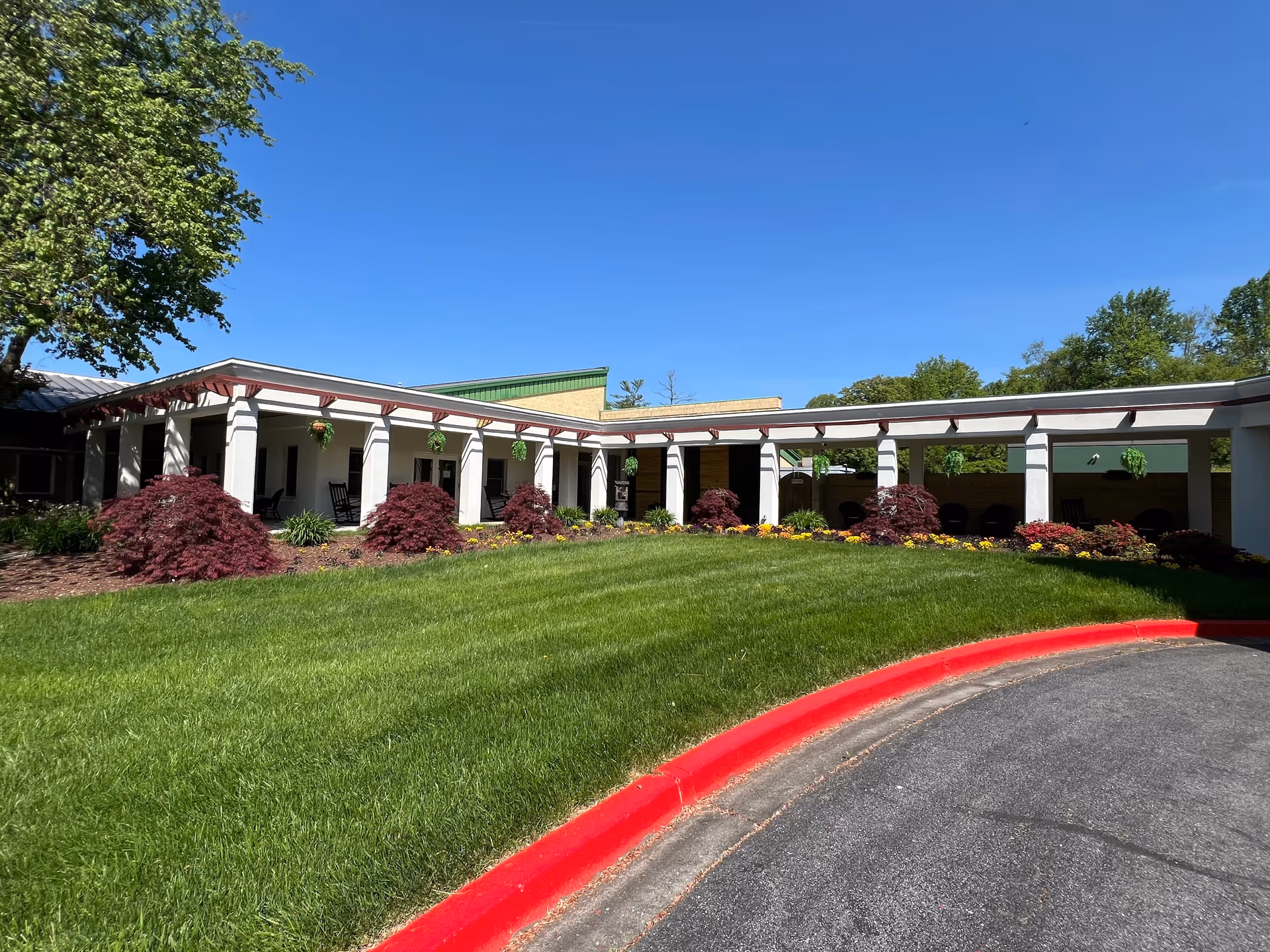 Exterior view of Regency Park Senior Living Community showing a single-story building with a covered walkway supported by white columns. The building is surrounded by a well-maintained lawn, landscaped bushes, and hanging plants under a clear blue sky.