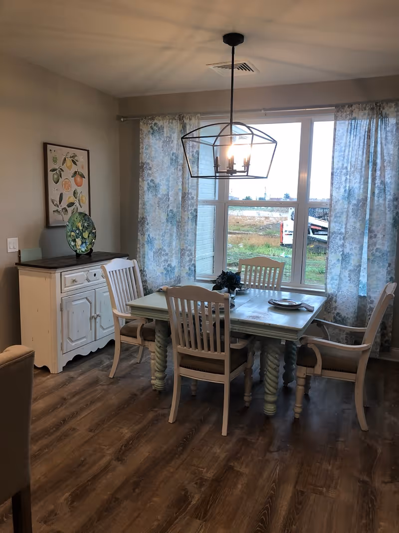 Dining room with a wooden table and chairs under a pendant light, a sideboard, and floral curtains covering large windows.