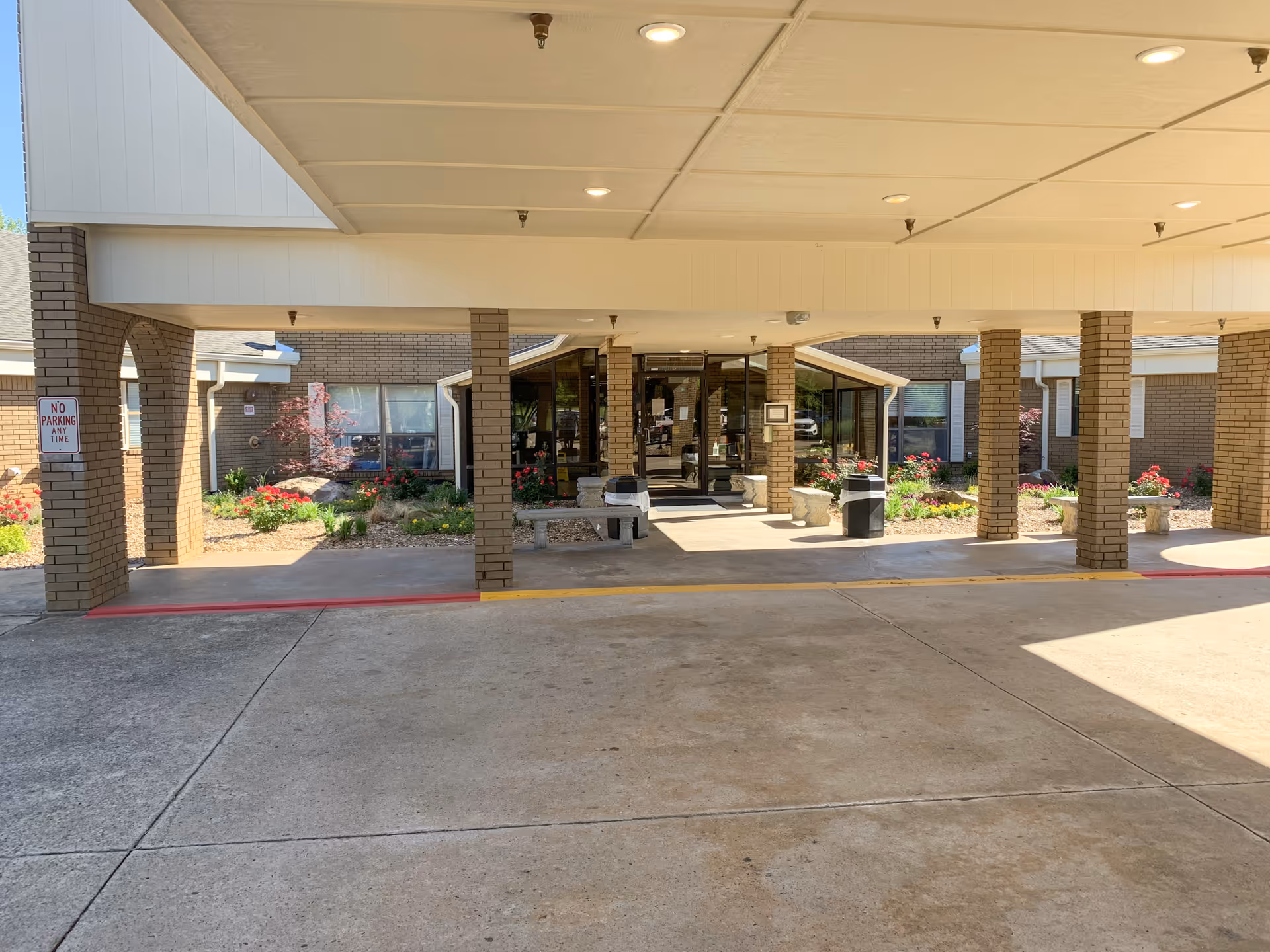 Covered entrance area of Oaks Healthcare Center with brick pillars, a concrete driveway, and landscaped flower beds. The entrance has glass doors and windows, with benches and a trash can nearby. A 'No Parking Any Time' sign is visible on one of the pillars.