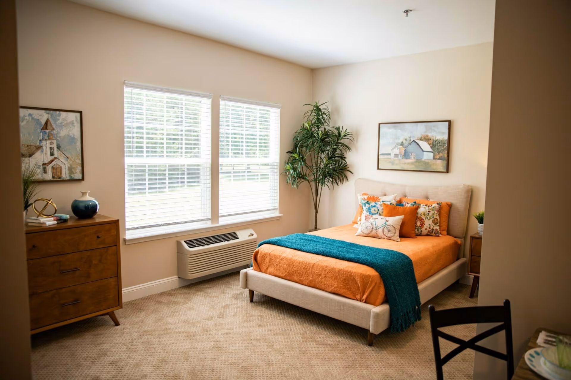 Bright furnished bedroom with an orange-and-teal bed, dresser, large window, and potted plant.