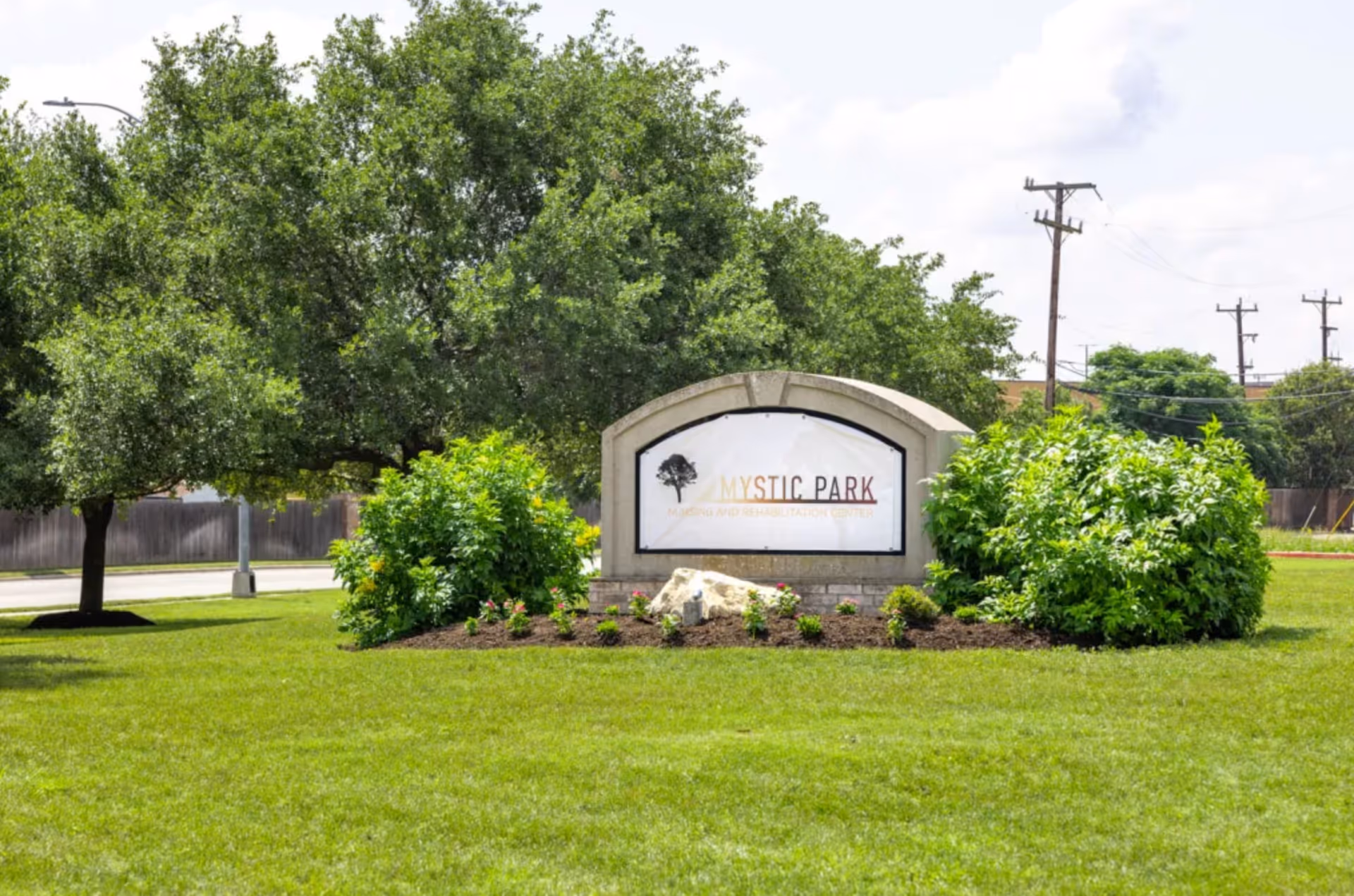 A landscaped outdoor area with green grass, bushes, and trees surrounding a stone sign that reads 'Mystic Park Nursing and Rehabilitation Center' under a partly cloudy sky.