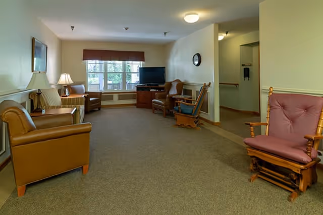 A spacious senior living common room with upholstered armchairs and wooden rocking chairs arranged around a television beneath a window.