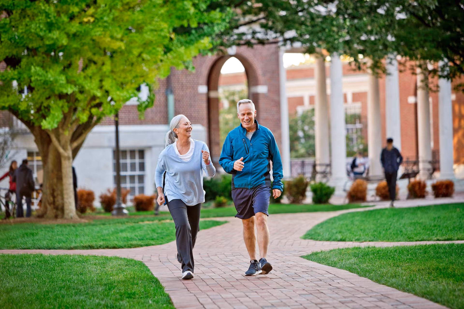 Two older adults jogging along a brick walkway on a leafy campus with a columned building in the background.