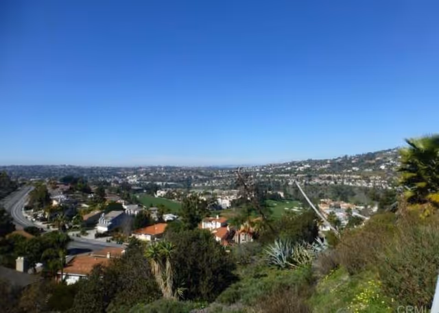 Panoramic view of a suburban area with houses, greenery, and hills under a clear blue sky.
