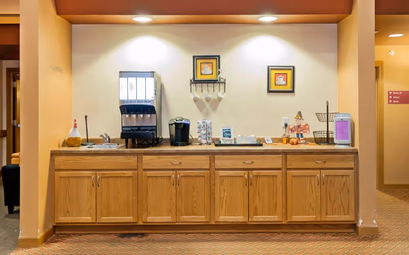 A beverage station in an assisted living facility with a wooden cabinet below and a countertop holding a soda fountain machine, a coffee maker, a stack of coffee pods, a small chalkboard sign, and a few decorative items including framed pictures of coffee cups on the wall above.