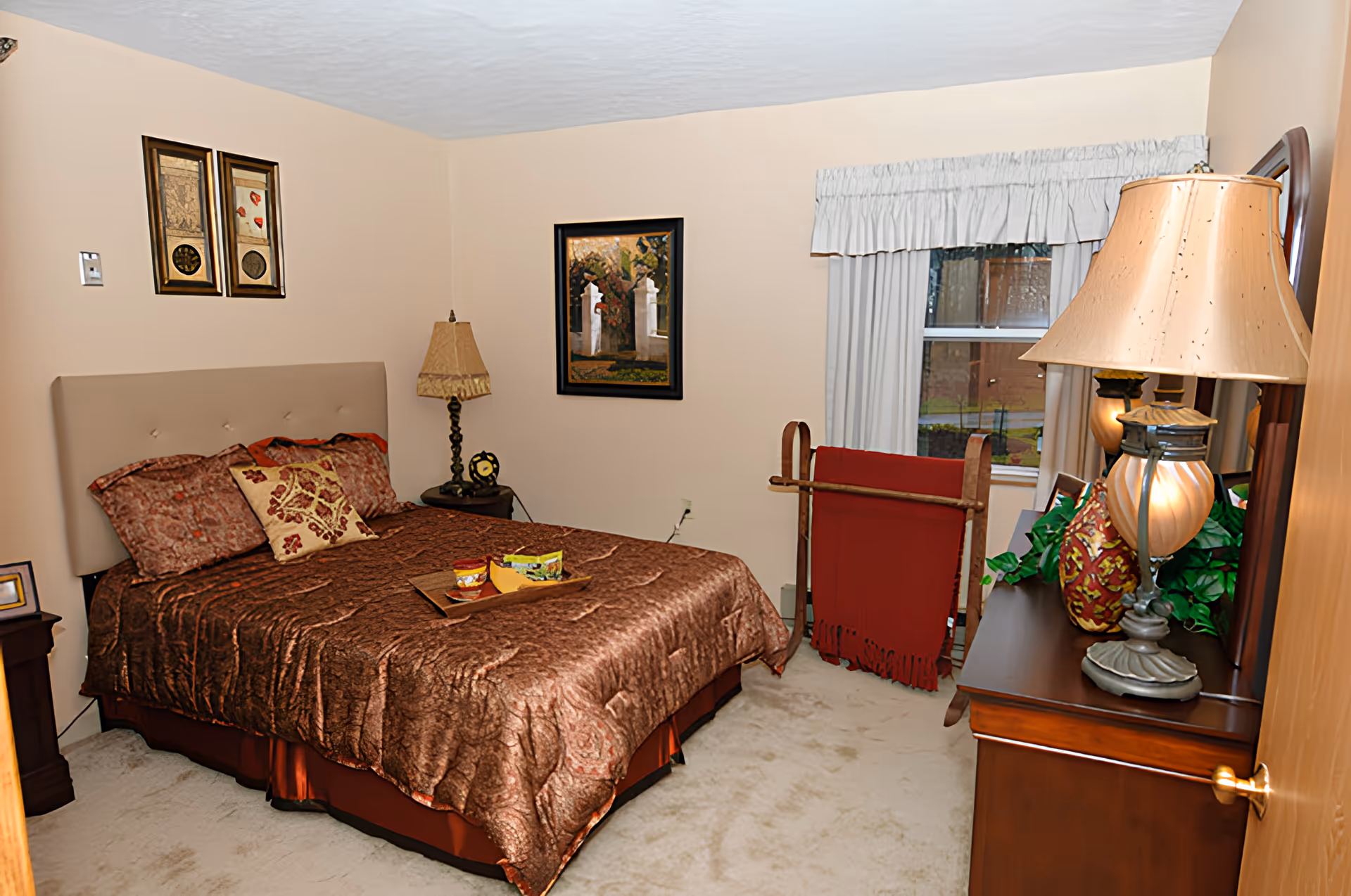 A cozy bedroom with a large bed covered in a brown patterned comforter and decorative pillows. There is a wooden nightstand with a lamp and a clock, a framed painting on the wall, a window with white curtains, and a wooden dresser with a lamp and decorative items. A red blanket is draped over a wooden rack near the window.