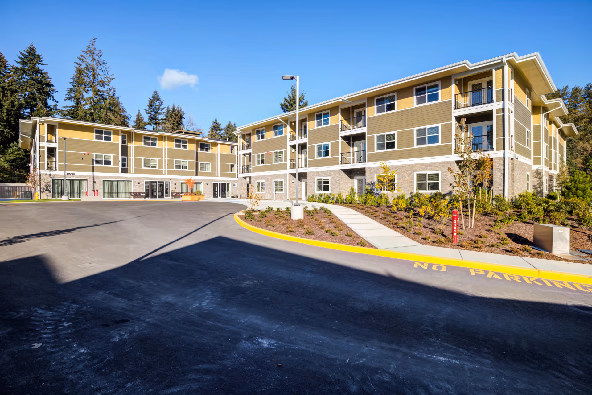 Exterior view of a modern three-story senior living community building with beige and yellow siding, multiple windows, balconies, and a paved driveway with a 'No Parking' zone. The building is surrounded by landscaped greenery and tall trees under a clear blue sky.