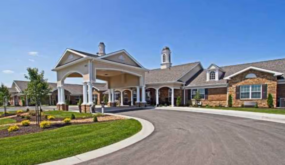 Exterior view of a single-story senior living facility building with a covered entrance supported by white columns, landscaped greenery, and a clear blue sky.