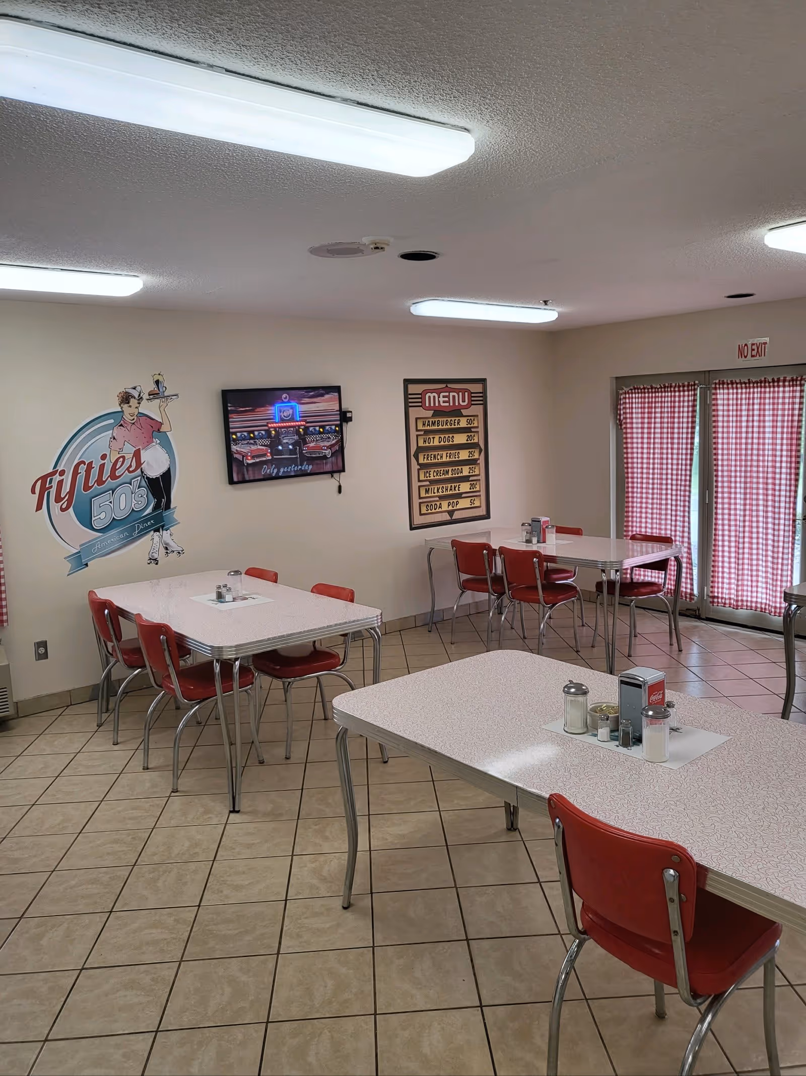 A retro-style dining room with red and white checkered curtains, red chairs, and white tables. The walls feature a vintage 'Fifties 50s' diner sign and a menu board listing items like hamburgers, hot dogs, french fries, ice cream soda, milkshake, and soda pop. The floor is tiled, and fluorescent lights illuminate the room.