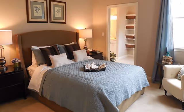 Neat furnished bedroom featuring a bed with a blue quilt, bedside tables and lamps, framed wall art, and an open doorway to a bathroom.