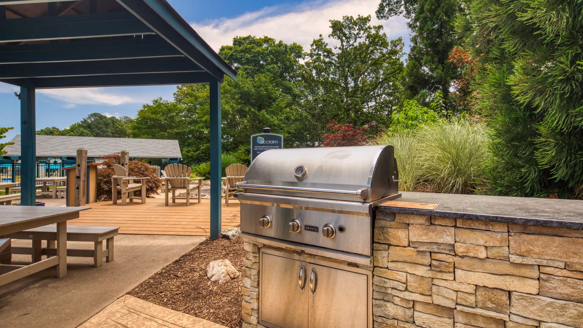 Outdoor patio area with a stainless steel built-in grill set into a stone countertop. There are wooden picnic tables under a blue pergola and Adirondack chairs on a wooden deck. The background features green trees and shrubs under a partly cloudy sky.