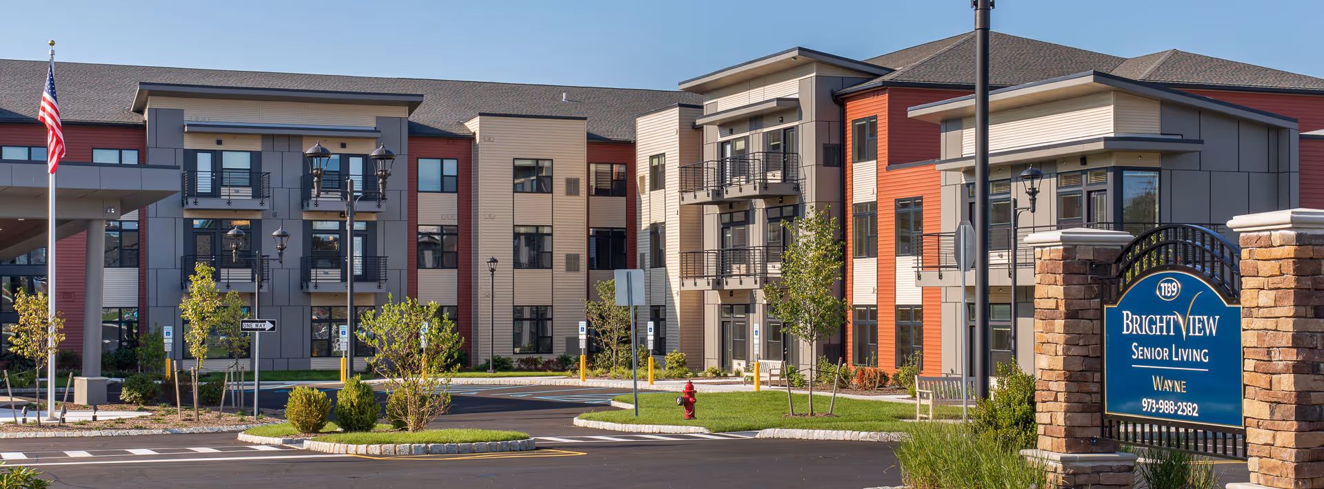 Exterior view of Brightview Wayne senior living facility showing a modern three-story building with balconies, landscaped greenery, and a sign at the entrance displaying the facility name and contact number.