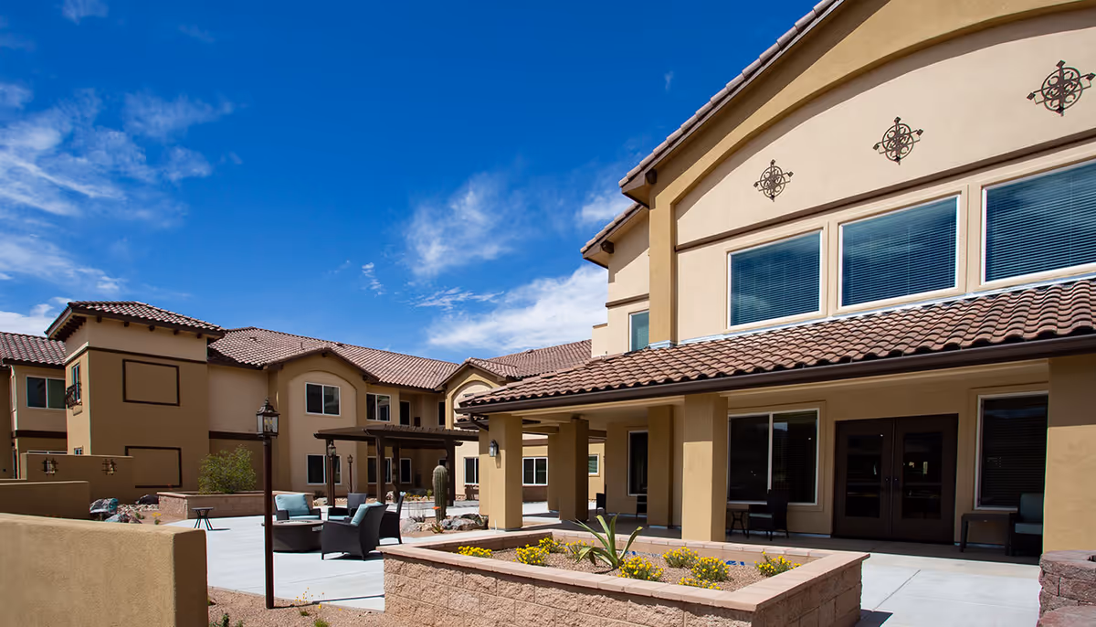 Exterior view of a senior living facility with beige stucco walls and red tile roofs under a blue sky. The courtyard features outdoor seating with chairs and tables, decorative plants, and a covered patio area.