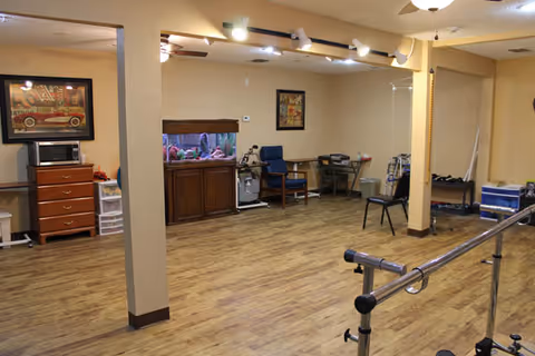 A spacious room with wooden flooring and beige walls, featuring exercise equipment such as parallel bars for walking support, a blue chair, a fish tank on a wooden cabinet, a microwave on a chest of drawers, and various small storage units. The room is well-lit with ceiling lights and has framed artwork on the walls.