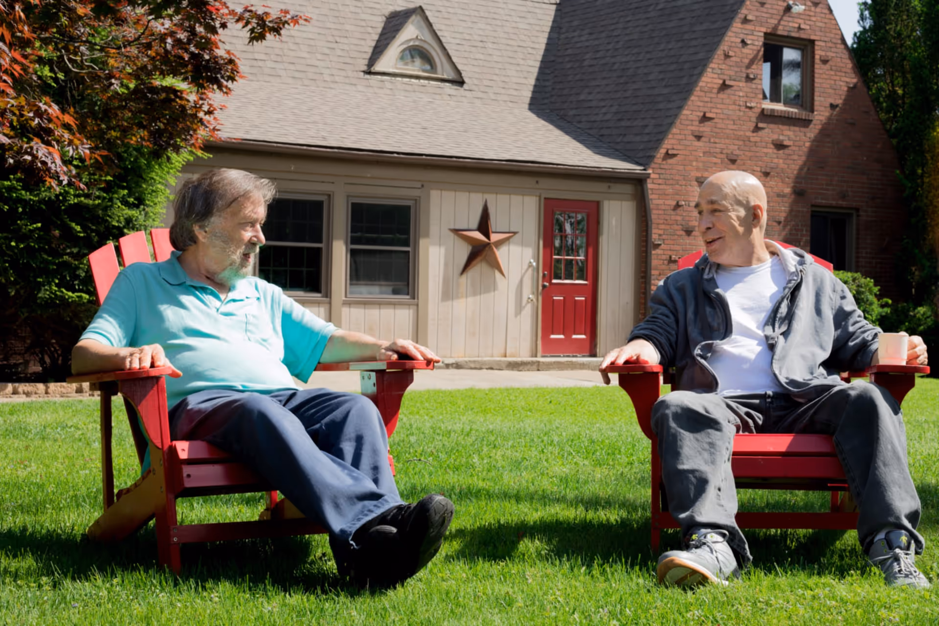Two elderly men sitting on red Adirondack chairs on a green lawn outside a house with a red door and a star decoration on the wall, engaged in conversation on a sunny day.