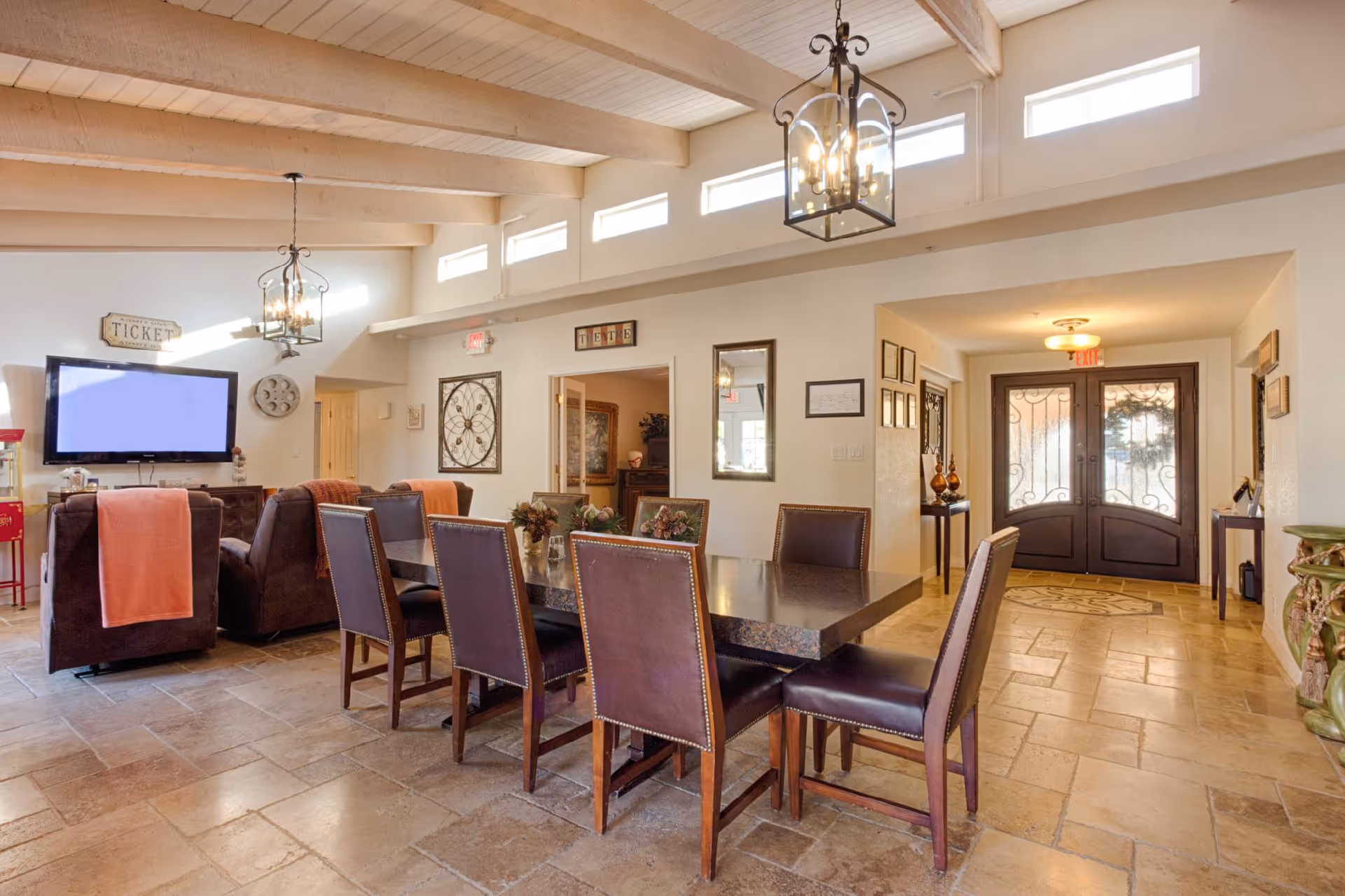 A spacious and well-lit common area with a large rectangular dining table surrounded by eight chairs. In the background, there are two comfortable armchairs with orange throws facing a wall-mounted TV. The room features high ceilings with exposed wooden beams and multiple small windows near the ceiling allowing natural light to enter. There are decorative wall hangings, two hanging lantern-style light fixtures, and double glass doors at the far end of the room.