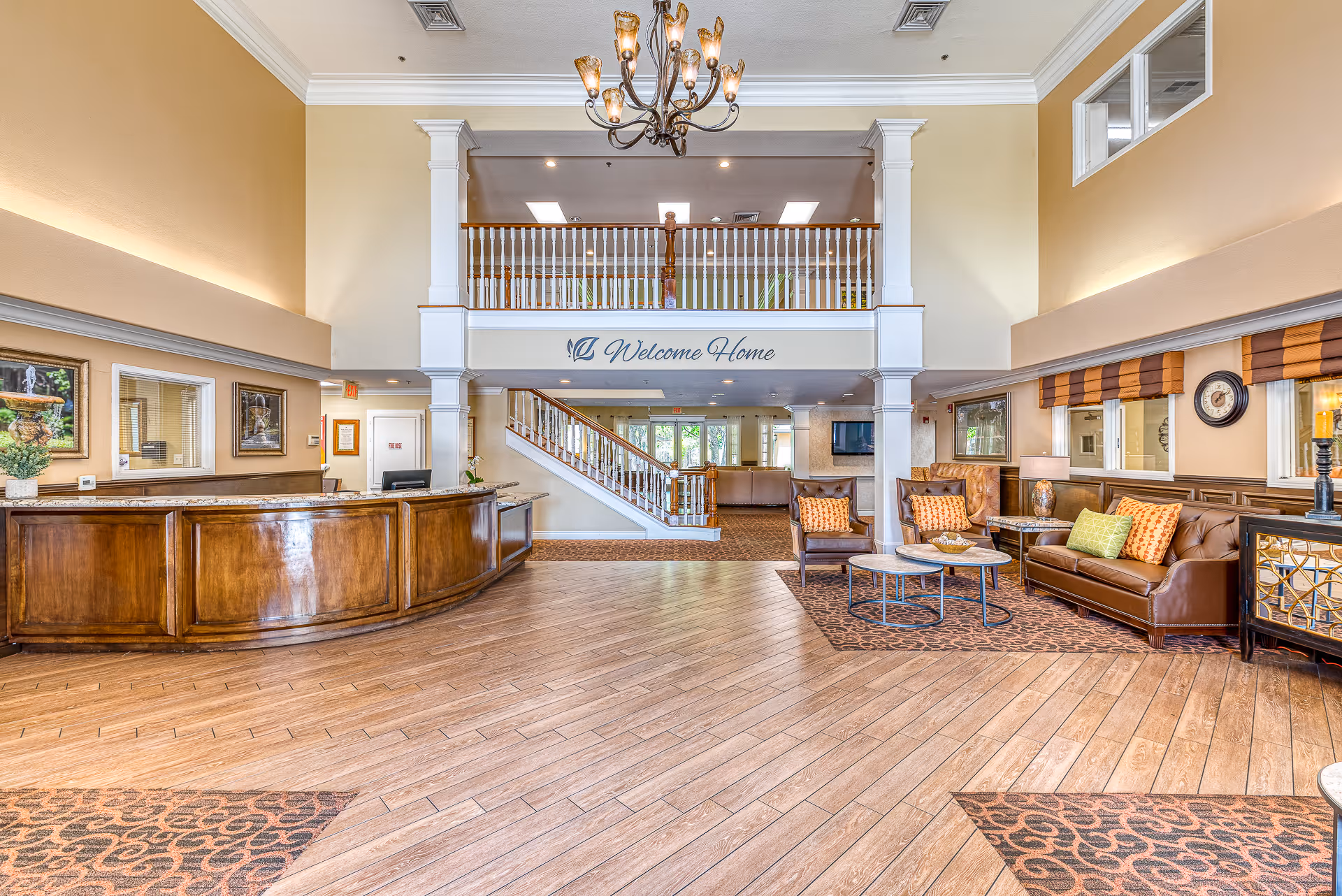 Spacious senior living lobby with a curved reception desk, seating area, chandelier, and a 'Welcome Home' sign above an upper balcony.