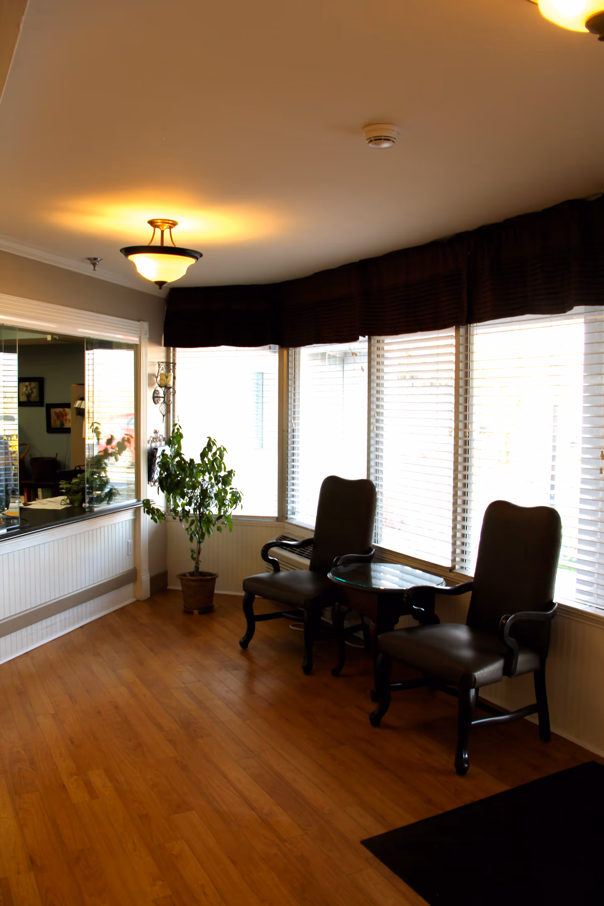 A cozy seating area with two dark wooden armchairs with cushions and a small glass-top table between them, placed near large windows with white blinds and dark valances. A potted plant is positioned near the corner, and the floor is wooden. The room is softly lit by ceiling lights.