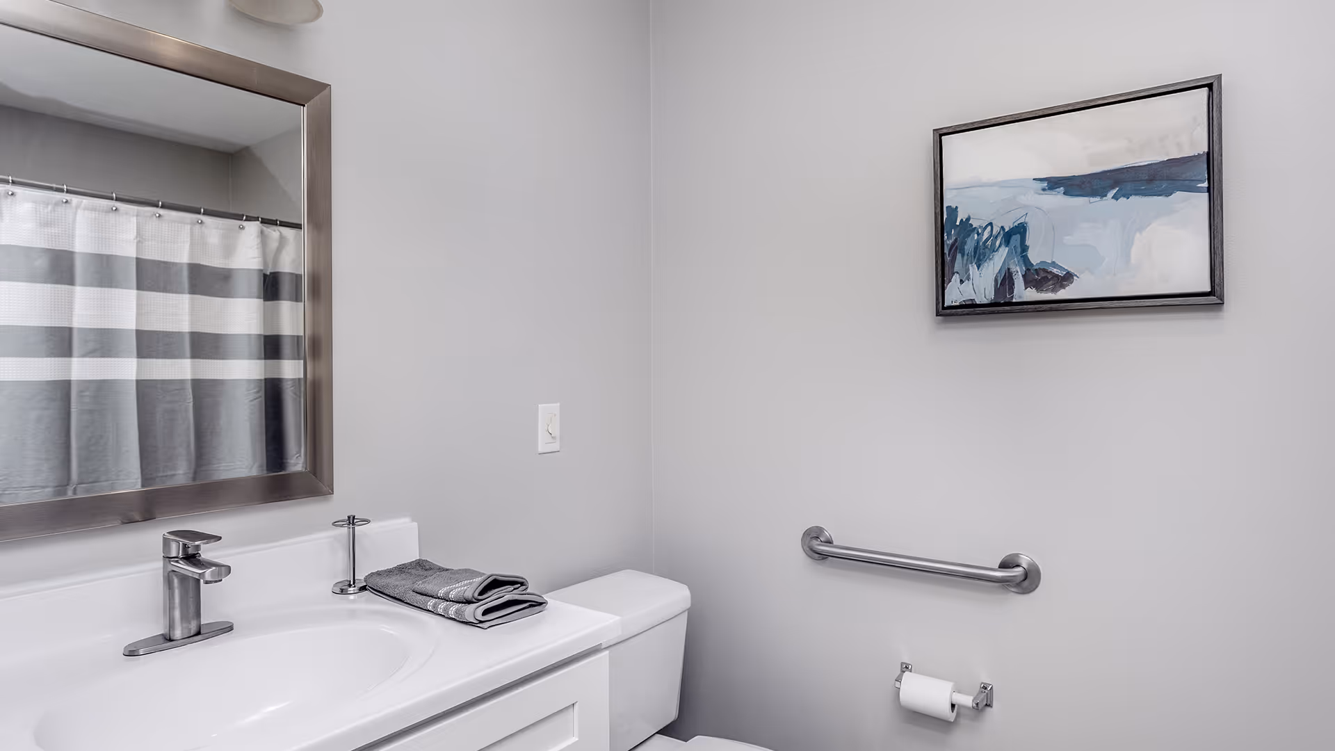 A clean bathroom with a white sink and countertop, a silver faucet, a large rectangular mirror with a silver frame, a toilet with a folded gray towel on top, a striped gray and white shower curtain, a metal grab bar on the wall, a toilet paper holder with a roll of toilet paper, and a framed abstract painting with blue and gray tones hanging above the grab bar.