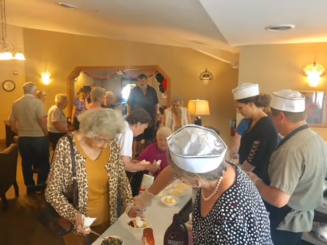 Elderly residents and staff in a communal dining room serving ice cream sundaes during a social event.