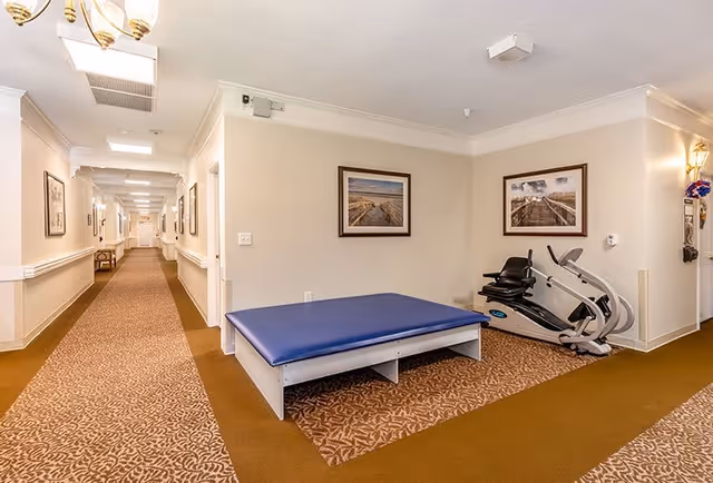 Interior hallway of a senior living facility with beige walls and patterned brown carpet. On the right side, there is a blue padded exercise mat and a recumbent exercise bike. The walls are decorated with framed landscape photographs. The hallway extends into the distance with doors and lighting fixtures along the walls.