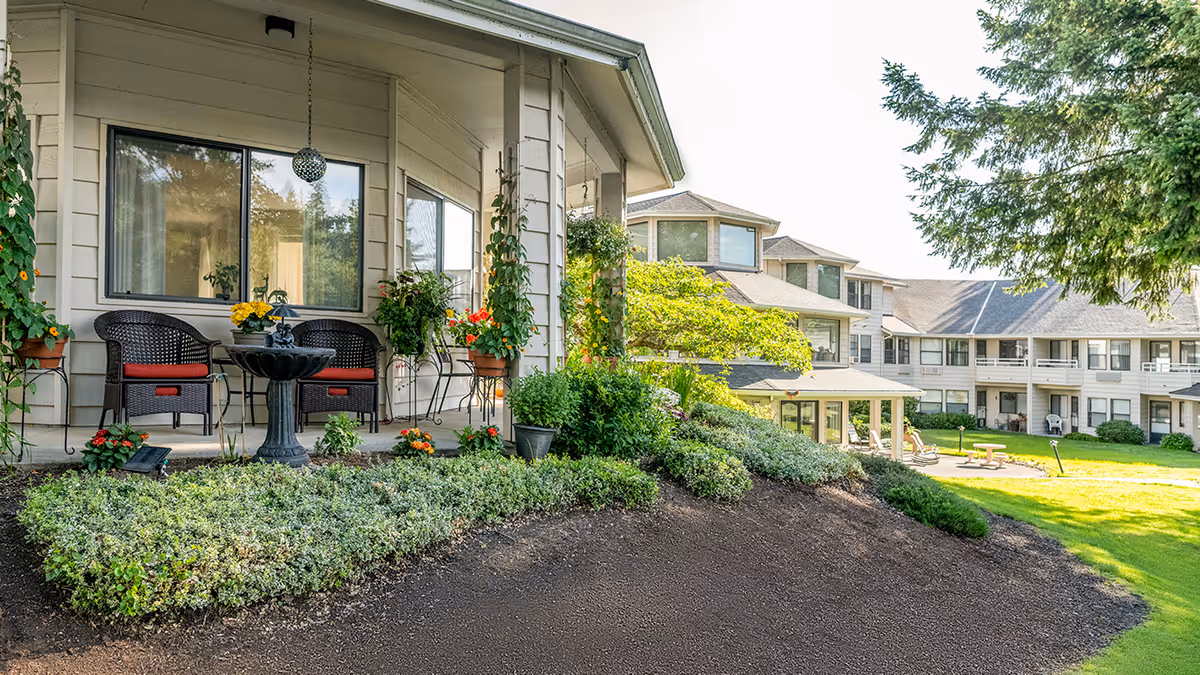 Outdoor patio area at Holiday Hidden Lakes with two wicker chairs with red cushions, a small black table with a flower pot, hanging plants, and well-maintained garden beds. In the background, there are multi-story residential buildings surrounded by green lawns and trees under a clear sky.